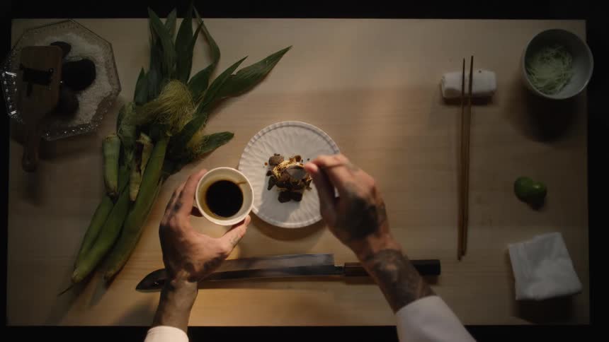 Overhead slow-motion view of a tattooed chef meticulously plating a gourmet meal, drizzling it with sauce