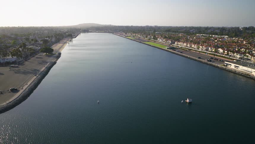 A static aerial view captures the tranquility of a wide coastal channel, flanked by residential homes and a road, under a clear sky