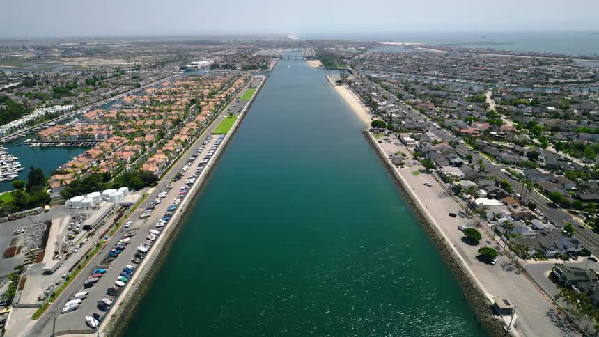 An expansive aerial view looks down a wide harbor channel flanked by marinas and waterfront homes, leading out to the distant ocean