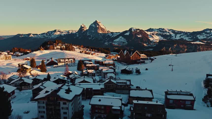 A stunning aerial view reveals a quaint snow-covered village in the Alps, bathed in the warm glow of sunrise