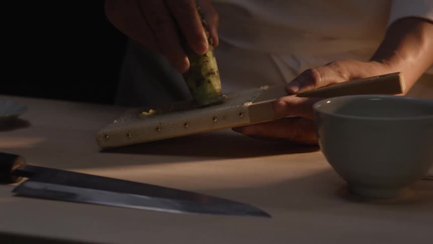 A sushi chef carefully grates a fresh wasabi root on a traditional sharkskin grater under a single spotlight