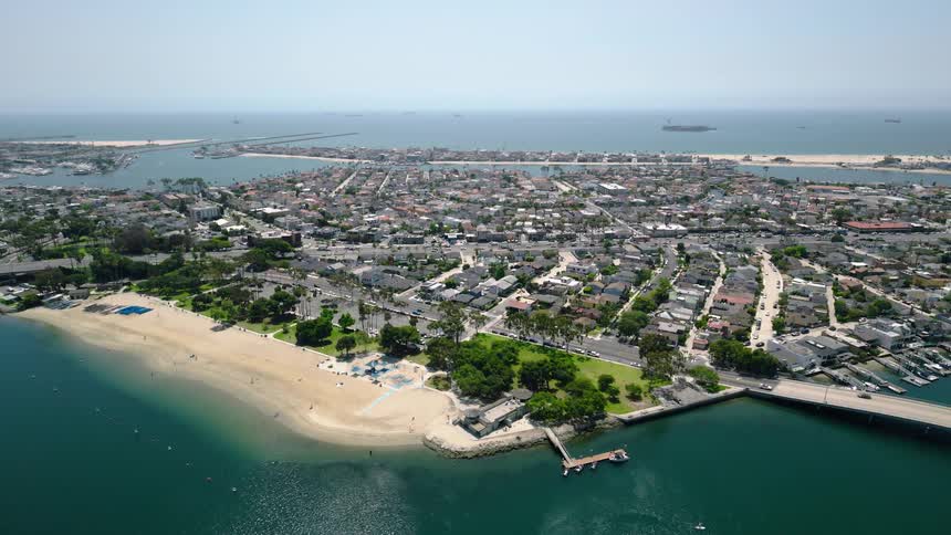 A slow aerial pullback shot reveals a beautiful coastal community in Southern California, showcasing the sandy beach, bay, bridge, and the ocean on a clear day