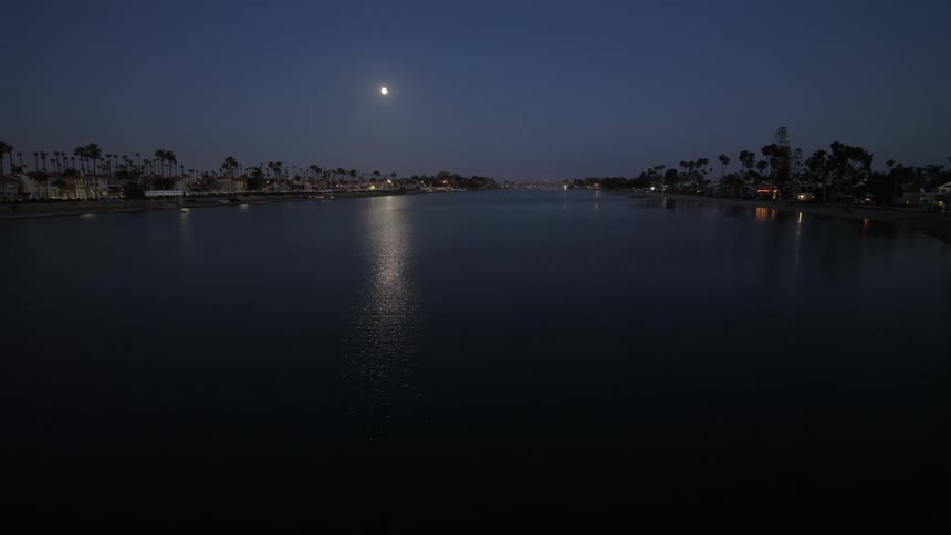 A slow pull-back aerial shot captures the peaceful ambiance of a coastal bay under a full moon at night