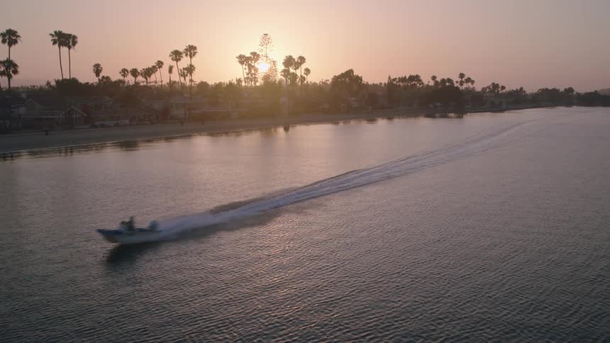 An aerial view captures a motorboat cruising across a tranquil harbor as the sun sets behind palm trees