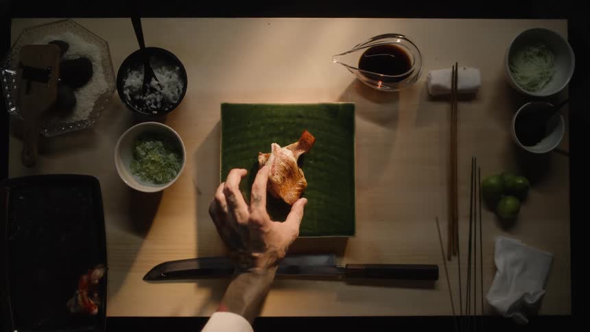 An overhead slow-motion cinemagraph captures a tattooed chef's hands artfully plating a piece of seared fish for a gourmet Japanese meal