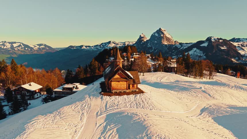 Golden sunrise light illuminates a secluded wooden chapel and village nestled among the snow-covered Swiss Alps in a stunning aerial view