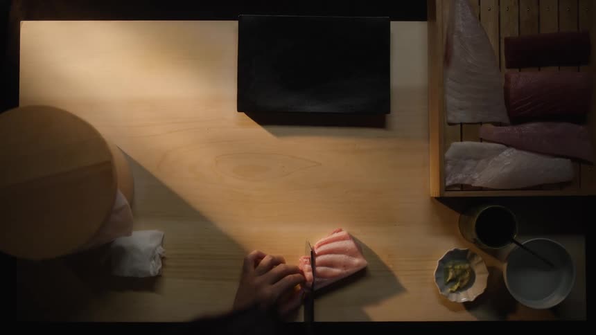 From a top-down perspective, a skilled sushi chef meticulously slices a fresh piece of raw fish for sashimi on a wooden cutting board