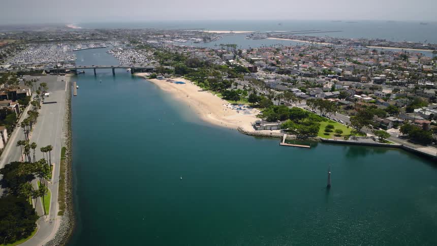 A stunning aerial view captures a picturesque coastal city with a calm bay, sandy beach, and bustling marina on a sunny day