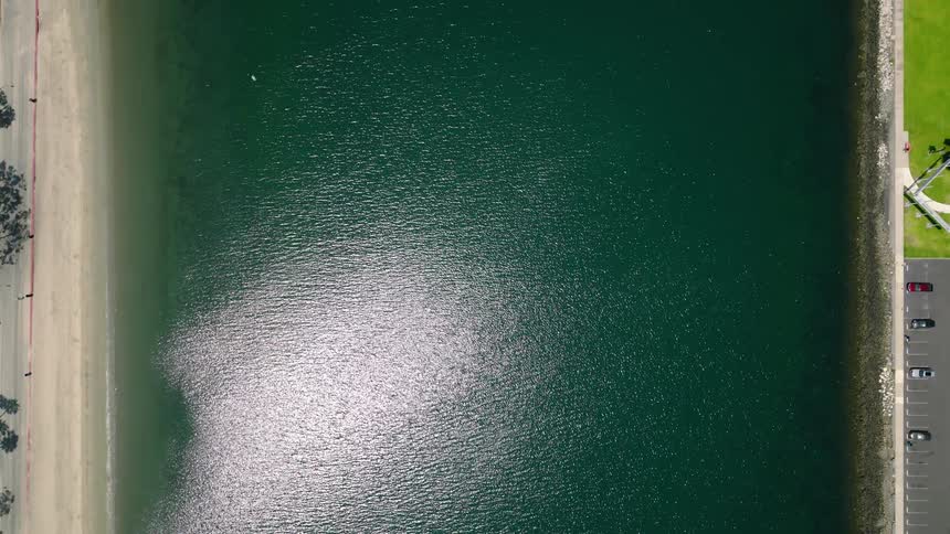 An aerial top-down view shows the sun glittering on the calm green water of a bay, bordered by a sandy beach on one side and a park on the other
