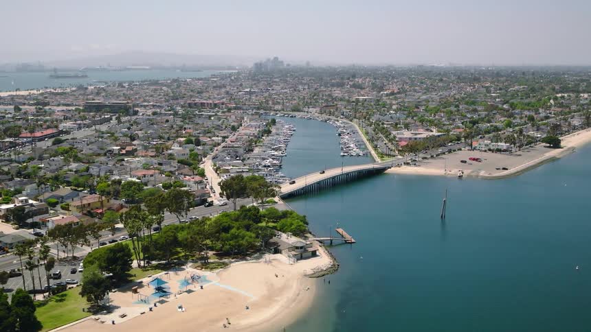 A slow-moving aerial shot captures a scenic coastal city with a sandy beach, a harbor full of boats, and a bridge connecting the neighborhoods on a sunny day