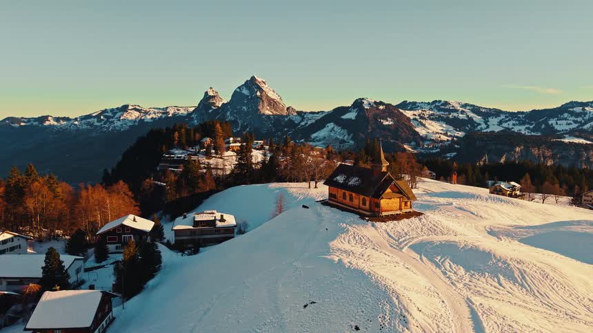 A drone flies over a serene snow-covered alpine village featuring a charming wooden church set against a dramatic mountain backdrop during golden hour