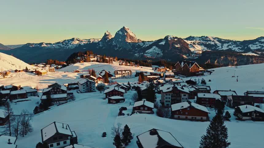 A stunning aerial shot pulls back over a quiet, snow-covered alpine village with majestic mountains basking in the golden sunrise