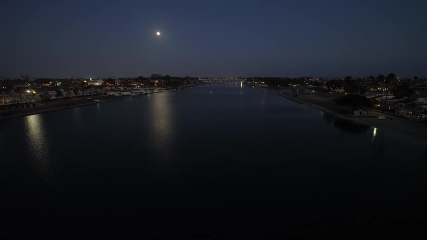Slow aerial dolly over a tranquil moonlit bay at night with the city lights of a coastal town glowing on the shorelines