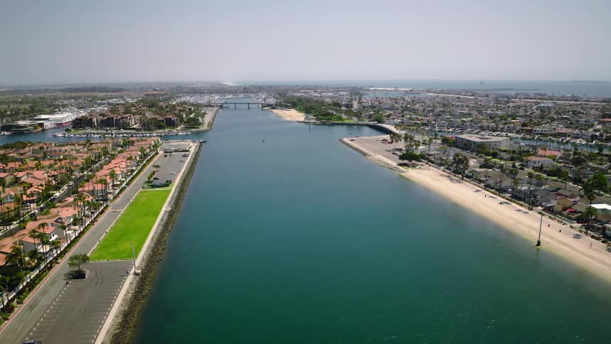 A slow, high-angle aerial view of a wide coastal channel, bordered by residential neighborhoods and a beach, with a marina and the ocean in the background