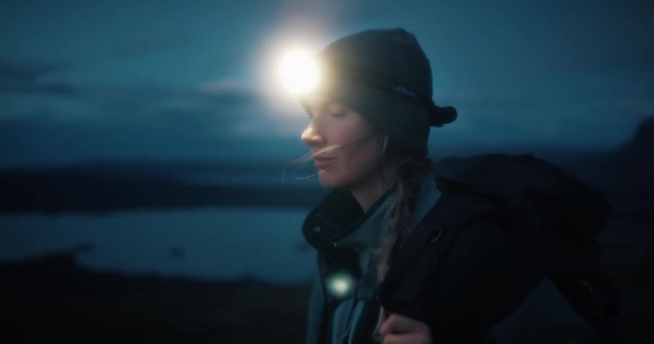 A female hiker wearing a backpack turns on her bright headlamp in the dark mountains at dusk in slow motion