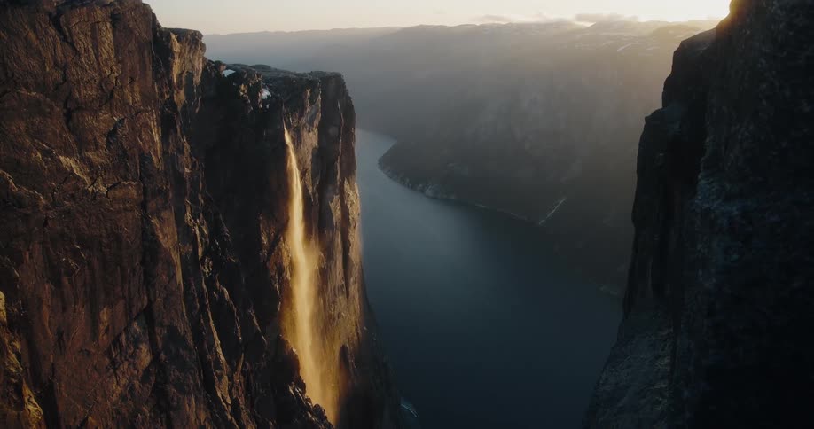 A majestic waterfall glows with the golden light of the setting sun as it cascades down a dramatic cliff face into a vast fjord