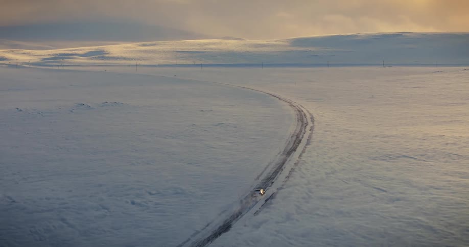 Aerial shot of a lone car driving down a remote, winding road through a vast, snow-covered landscape at sunset