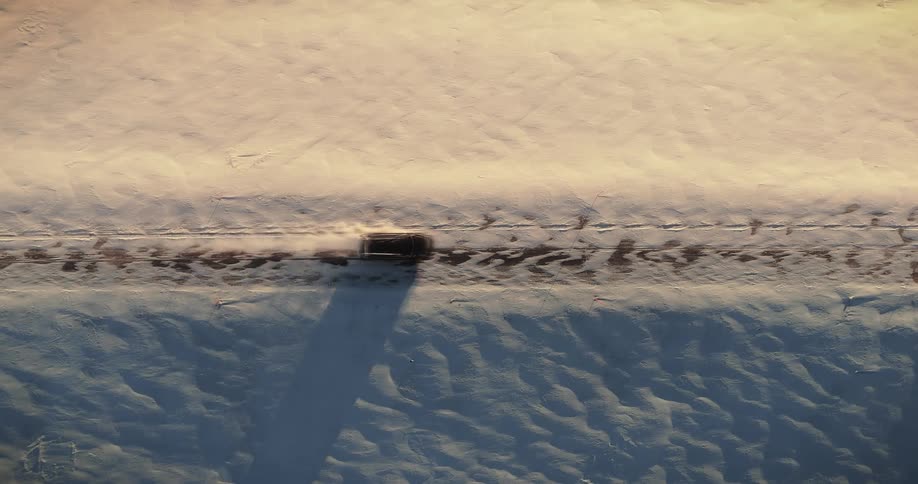 An aerial top-down view shows a car driving down a straight road through a vast, snowy landscape during golden hour