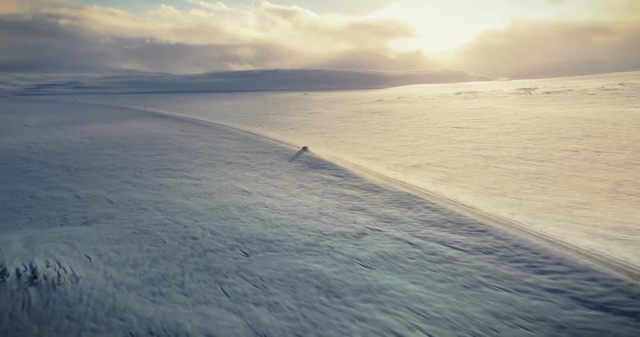 Epic aerial view of a single car driving along a remote road through a vast, snow-covered landscape at sunrise