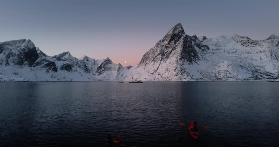 An aerial view shows two kayakers paddling across a serene fjord at dusk, with the moon rising over majestic, snow-covered mountains