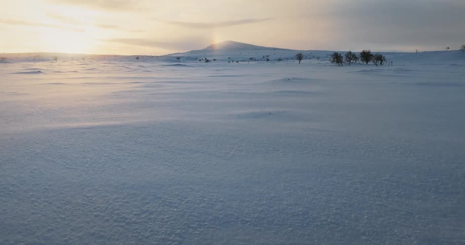 A low aerial shot glides over a vast, windy snowscape towards a distant hill during a beautiful winter sunset