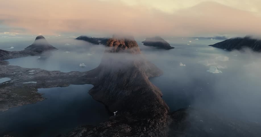 An epic aerial view captures fog clearing to reveal a dramatic mountain fjord landscape with icebergs in Greenland at sunrise