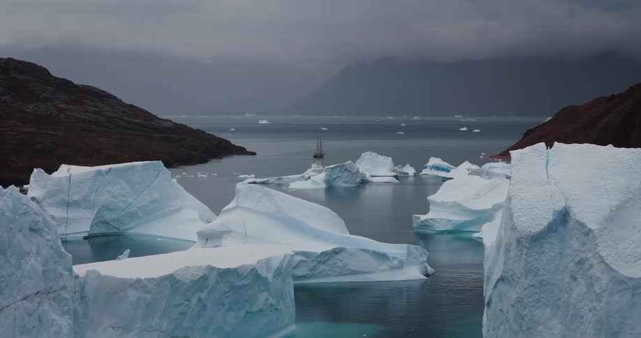 A solitary sailboat navigates through a massive ice field in a remote arctic fjord on a moody, overcast day