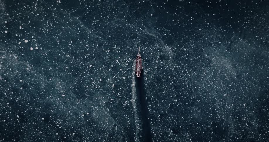 A solitary wooden sailing vessel rests in a vast, dark sea filled with drifting ice floes, captured in a dramatic top-down aerial cinemagraph