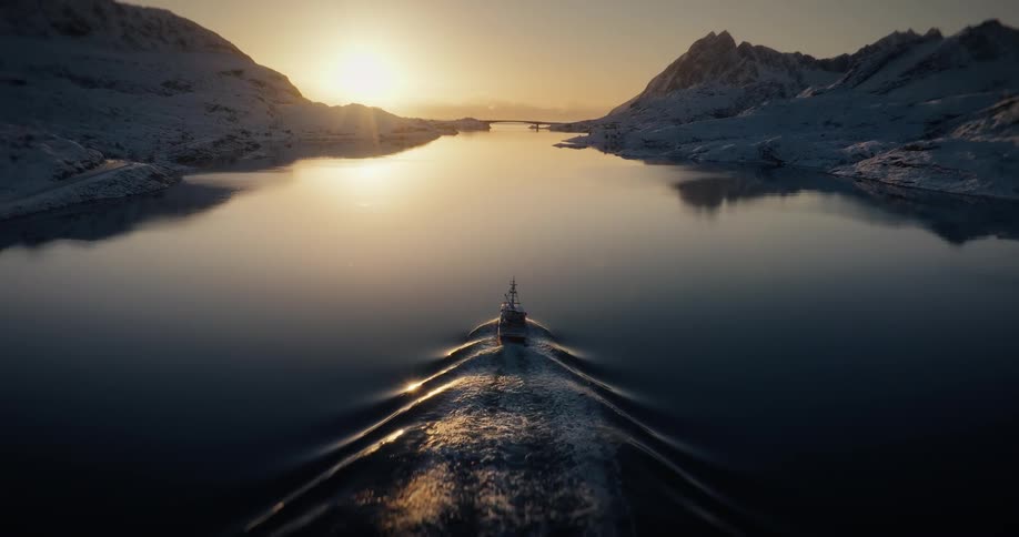 A serene aerial view of a boat sailing through a tranquil fjord between snow-covered mountains at sunrise, presented as a cinemagraph loop