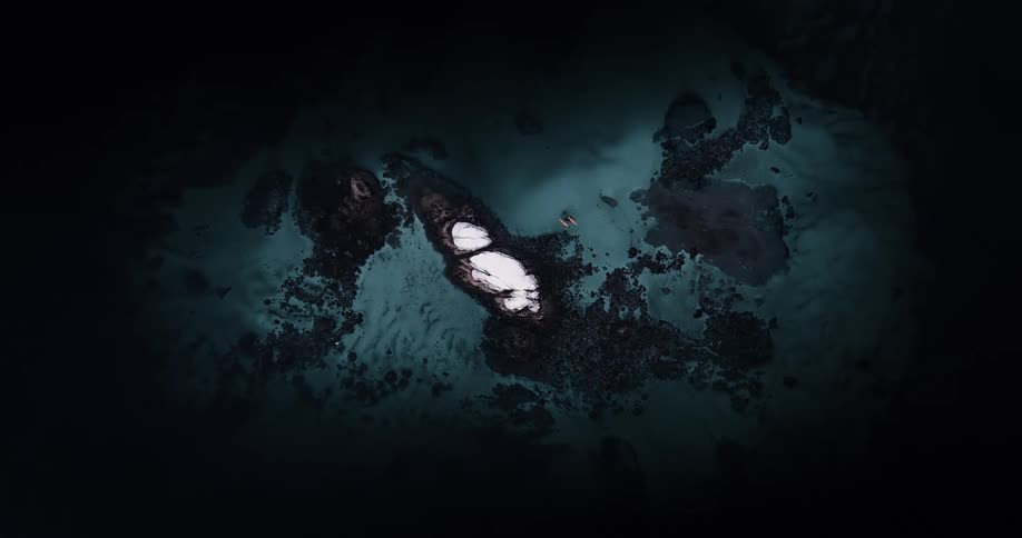 A dramatic top-down aerial shot follows two kayakers as they explore the dark, mysterious waters surrounding a small, rocky island