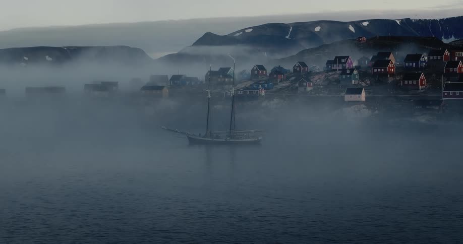 A traditional schooner is anchored in a misty bay with a colorful remote village on the shore
