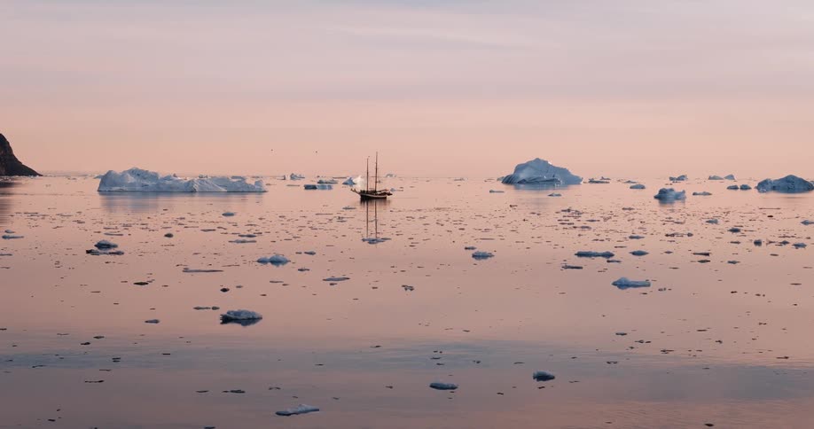 A vintage sailboat is anchored amongst floating icebergs in a calm arctic sea during a serene, pink-hued sunset