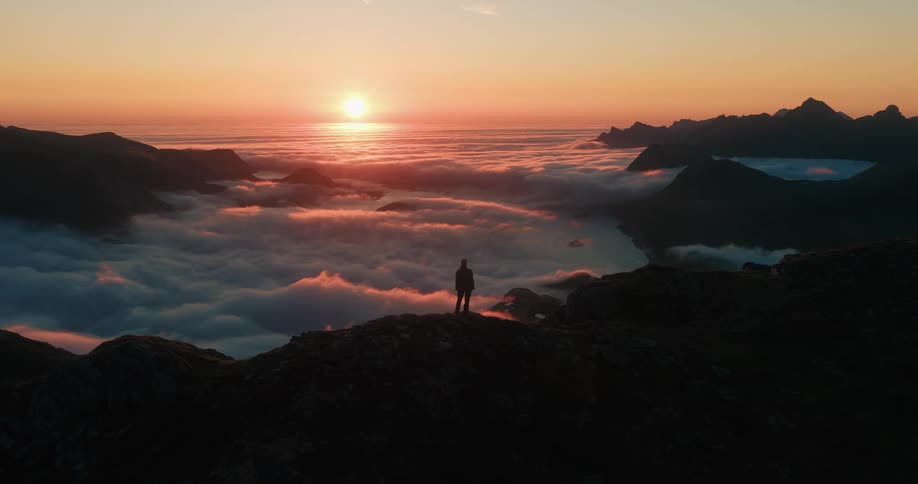 An aerial view pushes in on a lone hiker silhouetted on a mountain peak, watching an epic sunset over a sea of clouds covering the valley below
