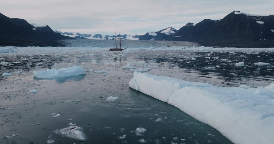 A classic sailboat is anchored in a serene arctic bay filled with floating icebergs in front of a massive glacier