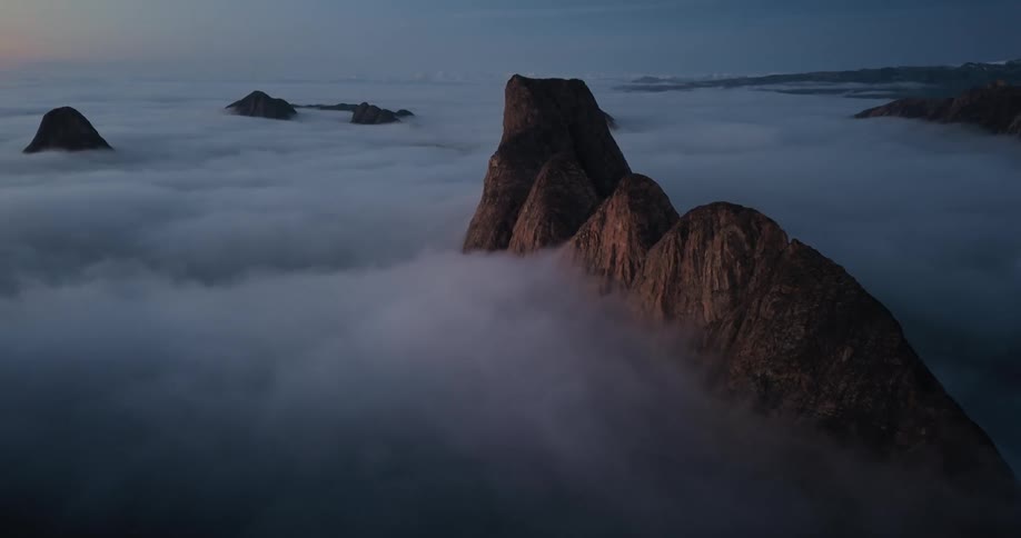 A slow pull-back aerial shot reveals a stunning mountain range piercing through a serene sea of clouds at dawn