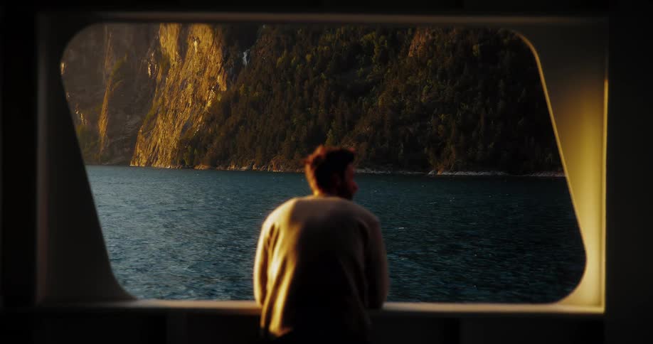 A silhouetted man looks out the window of a boat, admiring the majestic mountain fjords at golden hour