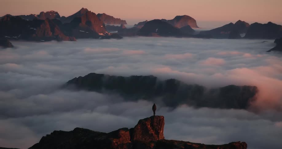 A solitary hiker stands on a mountain summit, gazing at a breathtaking sea of clouds filling the valleys below during a vibrant sunrise