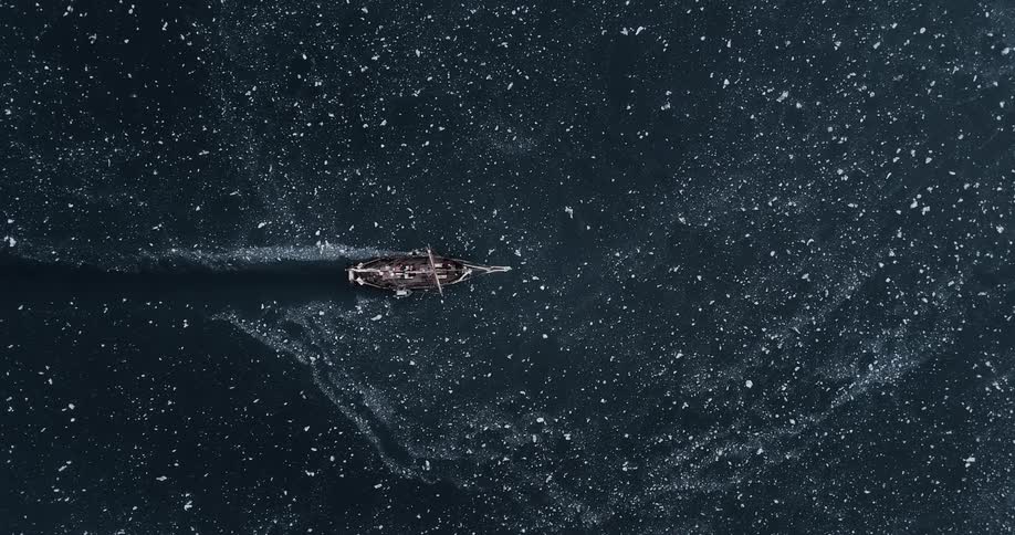 A top-down aerial cinemagraph shows an old wooden sailing vessel navigating through a dark, frozen sea