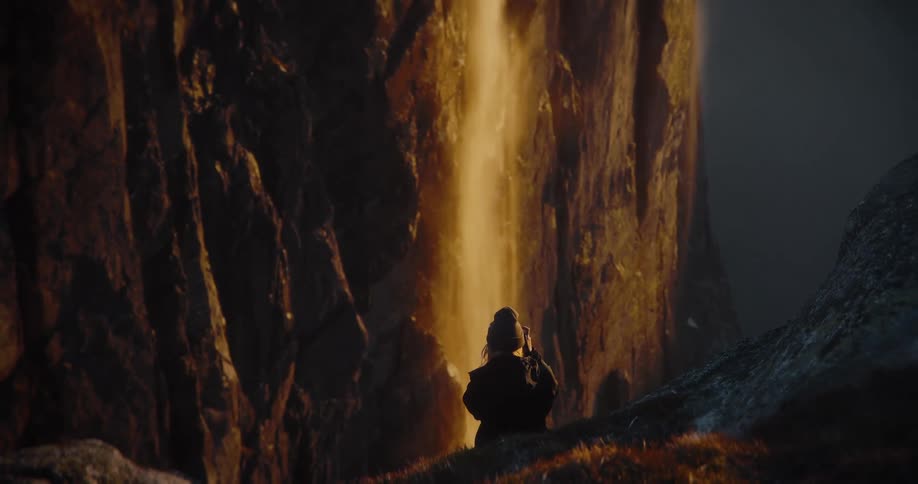 A lone figure sits watching a magnificent, glowing golden waterfall cascading down a dark mountain cliff