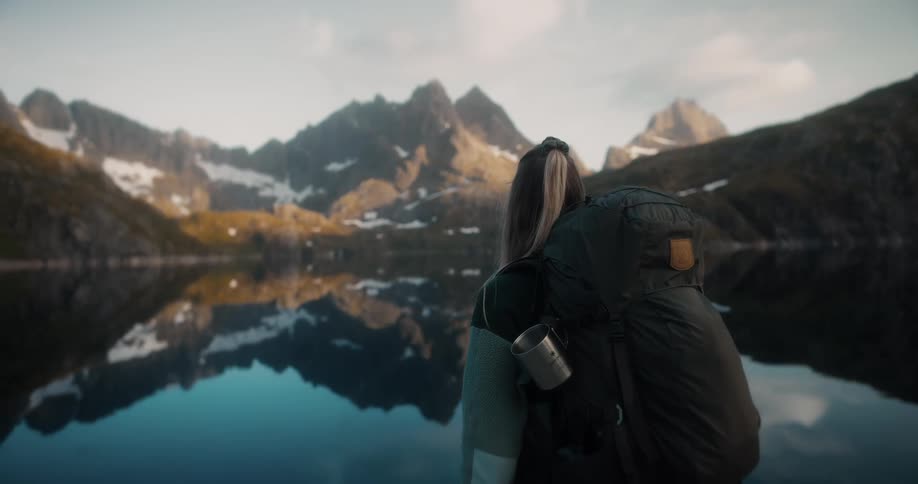 A female hiker with a large backpack peacefully gazes at the stunning reflection of snow-capped mountains in a tranquil alpine lake