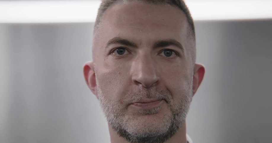 A close-up shot of a serious man with a beard and trendy haircut looking into the camera and then to the side