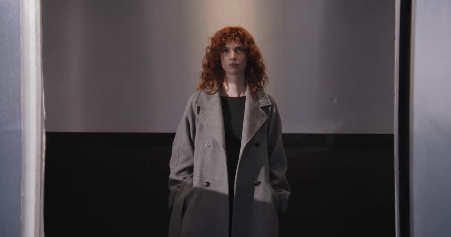 A young woman with curly red hair stands inside an elevator as the steel doors close in front of her