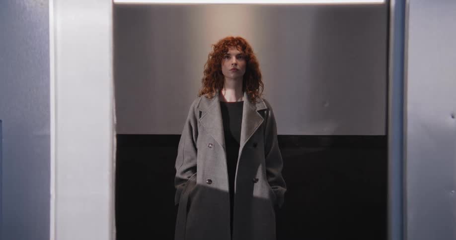 A young woman with curly red hair stands stoically inside an elevator as the shiny metal doors slowly close