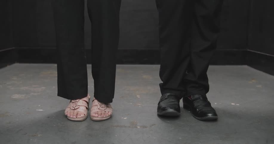 A low-angle shot of two pairs of feet, one fidgeting in sandals and the other still in dress shoes, waiting against a dark background