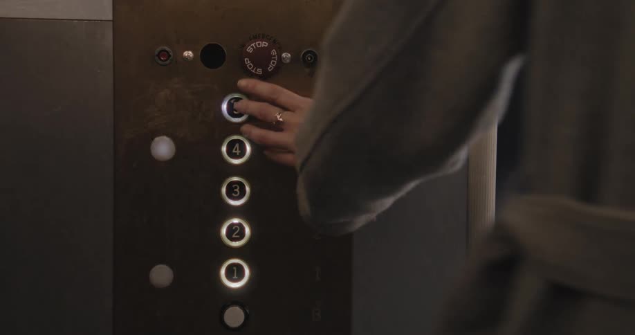 A woman's hand repeatedly presses the lit button for the fifth floor on a vintage elevator control panel as the doors close