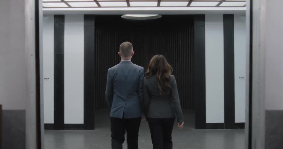 Rear view of a businessman and businesswoman walking out of an opening elevator into a stark, modern hallway