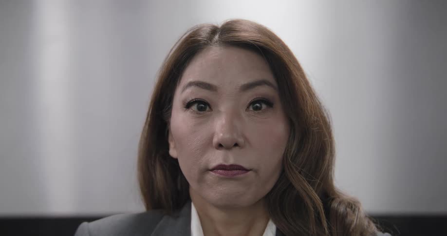 A close-up shot of an Asian businesswoman looking around nervously, then widening her eyes in shock as the camera slowly pulls back