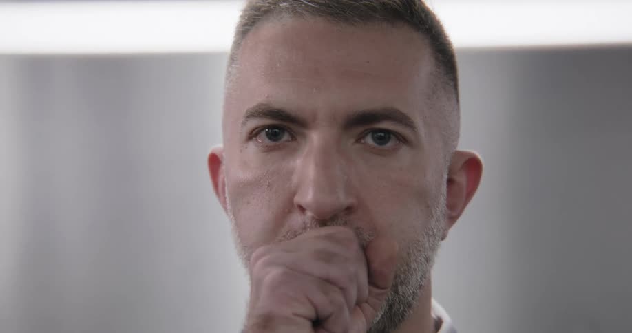 A close-up portrait of a serious man with blue eyes who looks worried and coughs into his fist