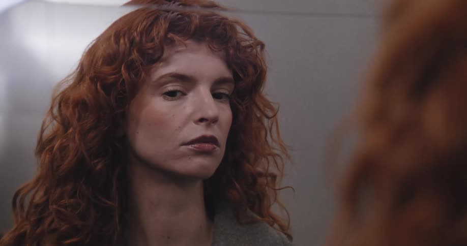 A contemplative red-haired woman with curly hair looks at her reflection in the mirror in slow motion