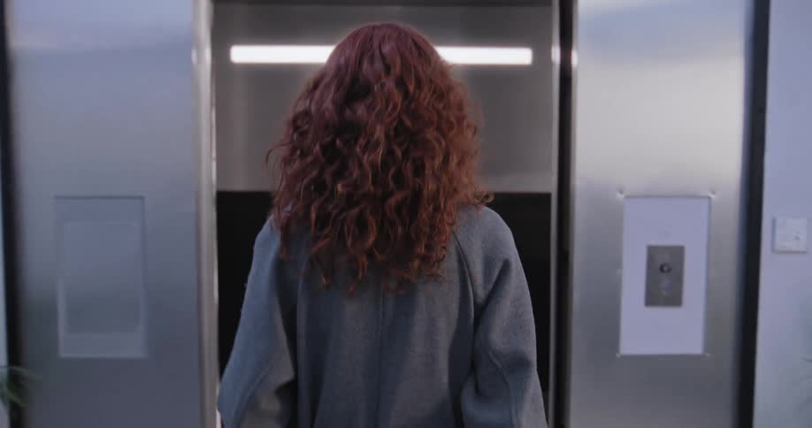 A stylish woman with vibrant curly red hair walks confidently into a modern elevator and turns to face the camera
